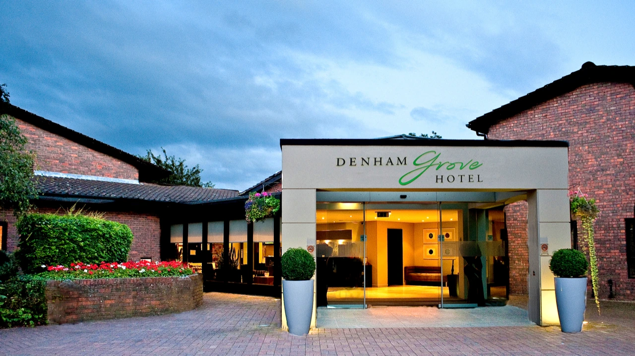 The main entrance of Denham Grove Hotel at dusk: a lit glass porch carrying the Denham Grove Hotel sign, clipped shrubs in tall planters either side, with the hotel's brick wings and a walled flower bed around it.