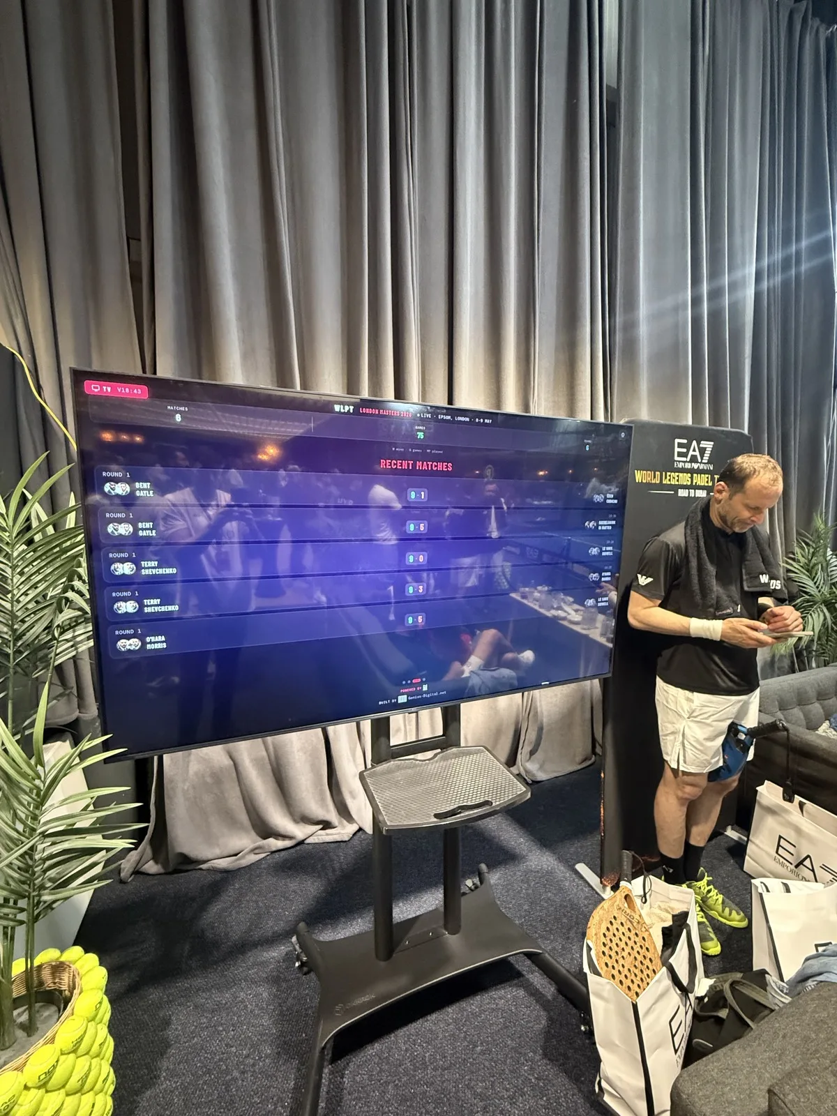 A courtside television on a stand showing the WLPT channel's recent matches feed, while a player in EA7 kit with a towel around his neck signs papers beside it.